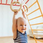 Cute baby performs gymnastic exercises on a wooden home sports complex stairs and rings.